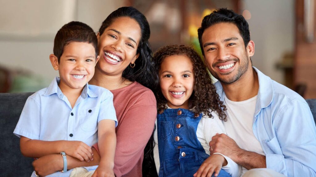 A mother, father, and two kids laughing together while taking a selfie at the kitchen table, representing conscious parenting and family connection on Mother's Day