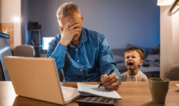 Busy mature businessman working from home and watching his crying son at night, who is is yelling at him. Upset displaced father forced to work from home as the pandemic corona virus (COVID-19) forces many employees to work from home. Real people. Copy space.